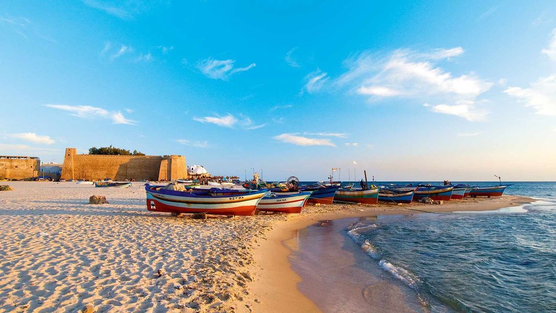 Des bateaux de pêche colorés sont alignés sur une plage de sable près de la mer, avec une grande forteresse en pierre en arrière-plan.