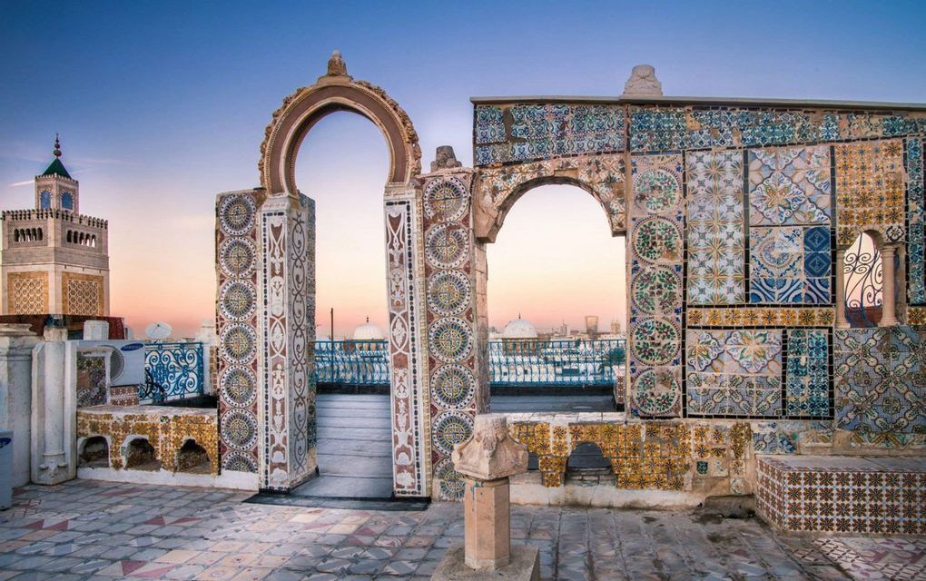 Une terrasse sur le toit avec des arches ornées et des mosaïques colorées, surplombant un paysage urbain avec un minaret au coucher du soleil.