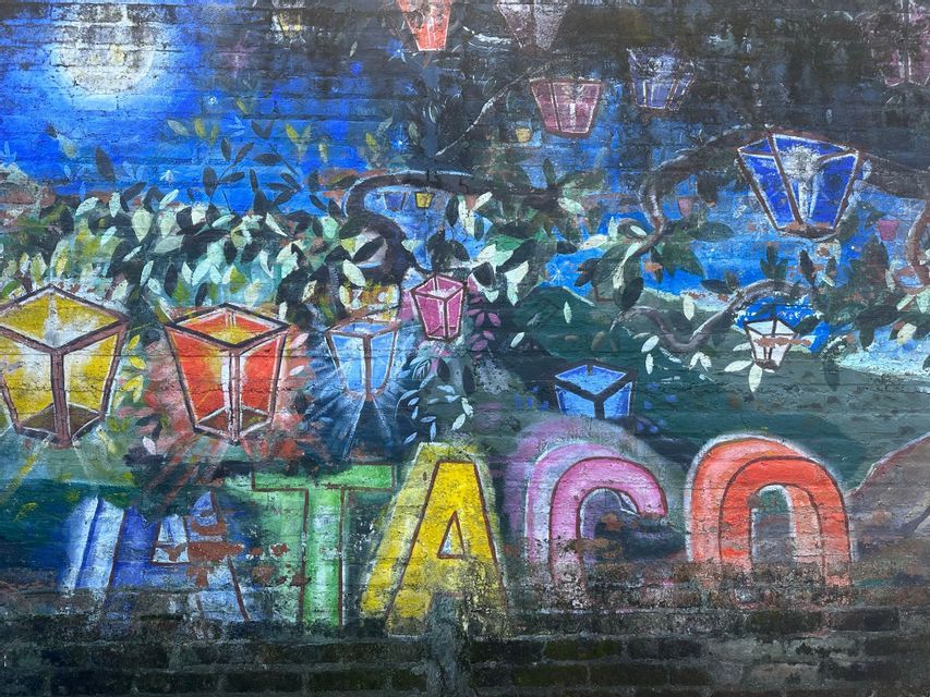 Ein farbenfrohes Graffiti-Wandbild auf einer Ziegelmauer, das einen Baum mit leuchtenden Laternen an den Ästen unter einem Vollmond zeigt.