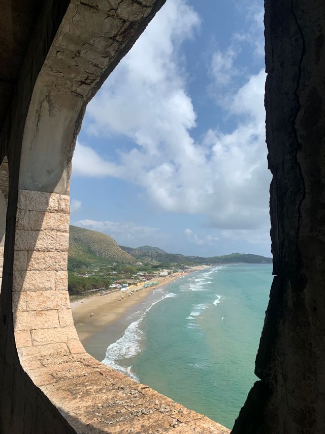 Una vista dall'interno di un arco in pietra mostra una lunga spiaggia sabbiosa, acqua turchese e verdi colline distanti sotto un cielo nuvoloso.