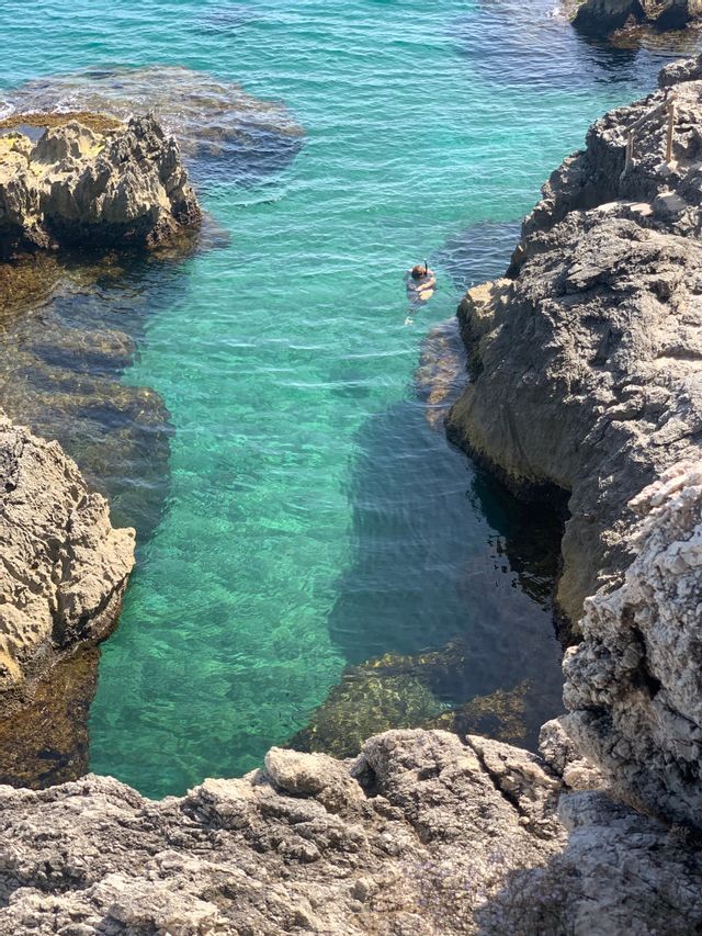 Vista dall'alto, una persona fa snorkeling in una cala rocciosa con acqua trasparente color turchese.