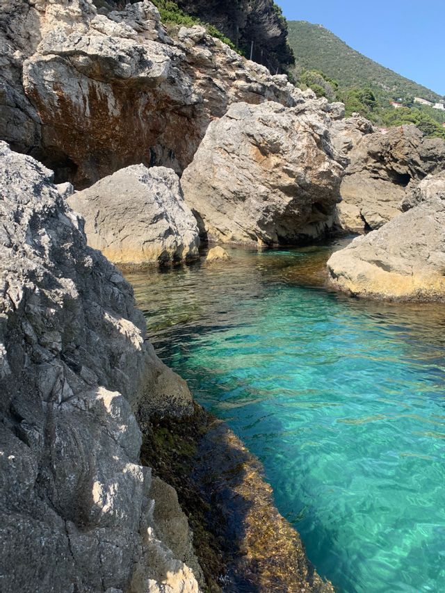 Acqua turchese limpida riempie una caletta rocciosa assolata, circondata da grandi formazioni rocciose grigie e un verde versante collinare.