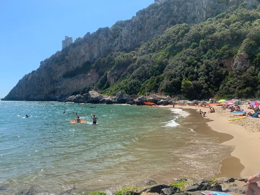 Le persone nuotano e si rilassano su una spiaggia sabbiosa ai piedi di una grande scogliera alberata, con una torre di pietra in cima.