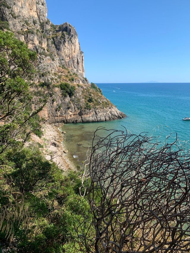 Una veduta dall'alto di una scogliera aspra e ricoperta di vegetazione che digrada verso una spiaggia di ciottoli appartata, bagnata da un mare calmo e turchese, sotto un cielo sereno.