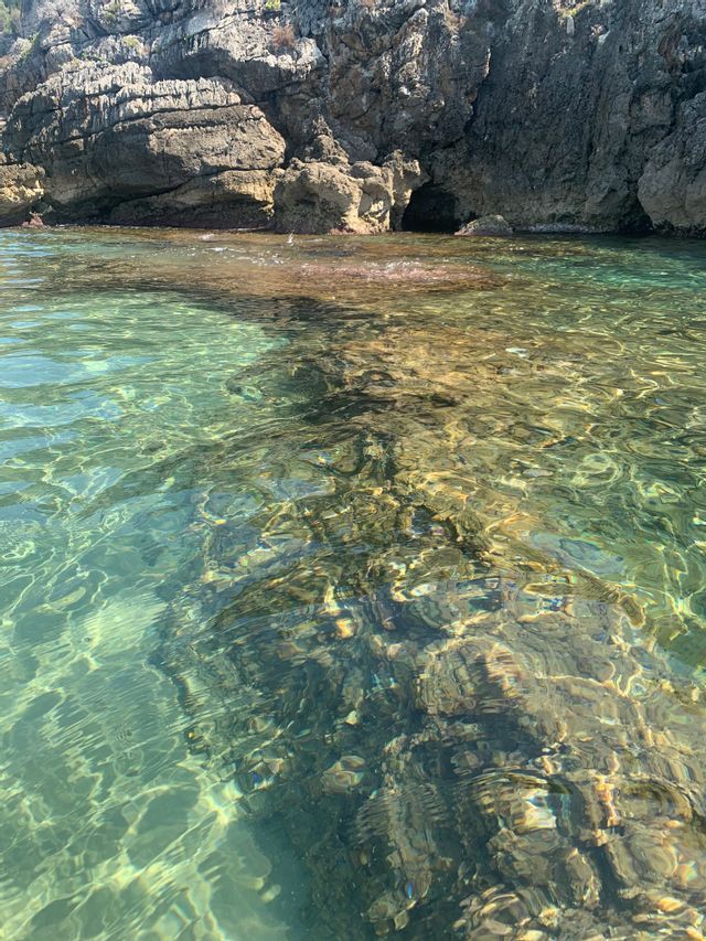 Acqua cristallina color turchese che si increspa su un fondale roccioso, con una scogliera impervia e una piccola grotta sullo sfondo.