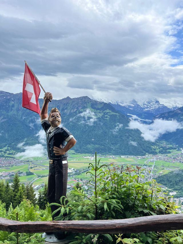 Une statue en bois d'un homme tenant un drapeau suisse surplombe une vallée verdoyante avec des montagnes enneigées en arrière-plan.