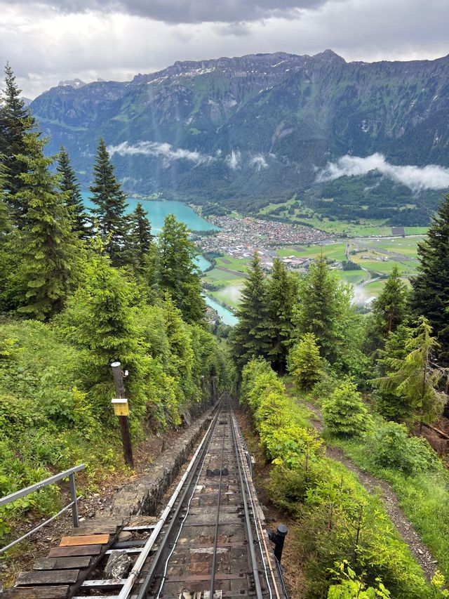 Un funiculaire escarpé descend une montagne boisée, offrant une vue sur un lac turquoise et une ville dans la vallée en contrebas.