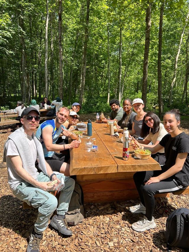 Un groupe WeRoad souriant pour une photo en mangeant à une table de pique-nique en bois dans une forêt ensoleillée.