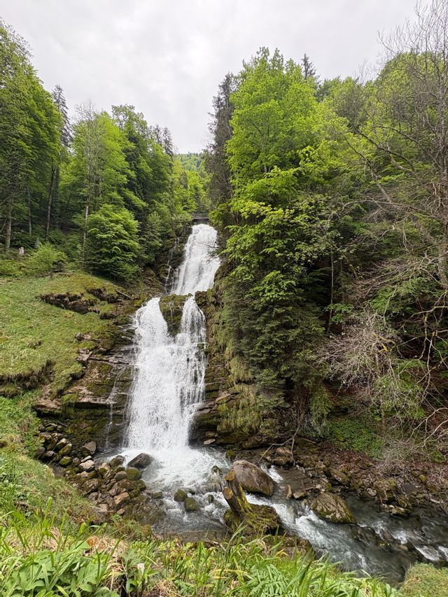 Une cascade à plusieurs niveaux dévale une falaise rocheuse, entourée d'une forêt verdoyante sous un ciel nuageux.