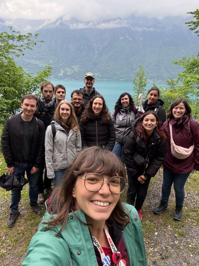Un groupe WeRoad prend un selfie sur un sentier surplombant un lac turquoise et des montagnes boisées sous un ciel nuageux.