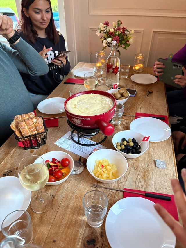 Un voyage de groupe WeRoad déguste une fondue au fromage à une table en bois garnie de pain, d'olives, de légumes et de vin.