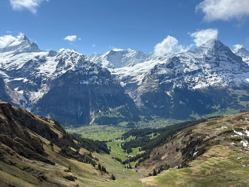 Une vue plongeante sur une vallée verdoyante nichée entre les montagnes, avec une chaîne de montagnes enneigée imposante à l'arrière-plan sous un ciel bleu.