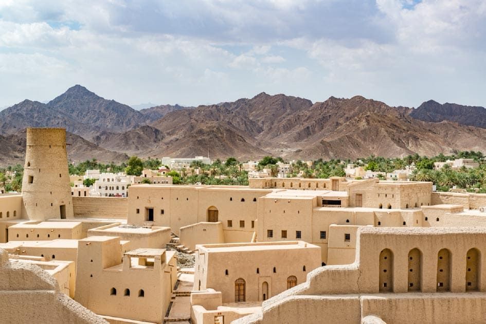 A historic village of sand-colored buildings and a round tower, with a green oasis and rugged mountains in the background.