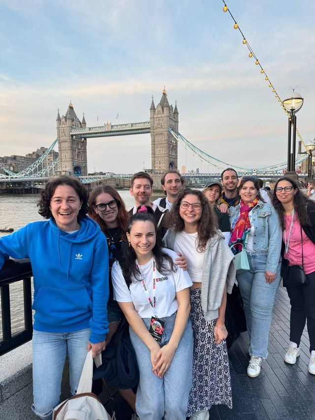 Un gruppo WeRoad in viaggio posa sorridendo per una foto su un lungomare, con un grande ponte storico visibile oltre il fiume.