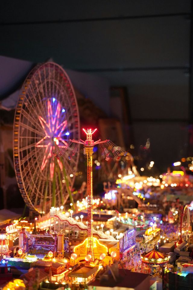 Maqueta en miniatura de un parque de atracciones iluminado de noche, con noria y columpio gigante en primer plano.