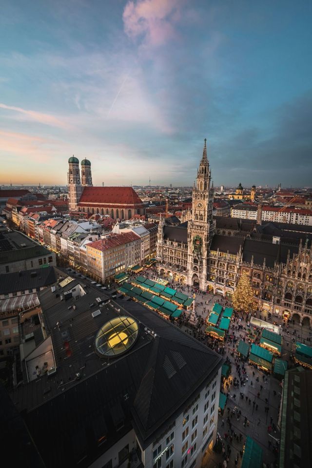 Una vista aérea de un animado mercado navideño en una plaza de la ciudad al atardecer, con puestos, multitudes y arquitectura histórica.