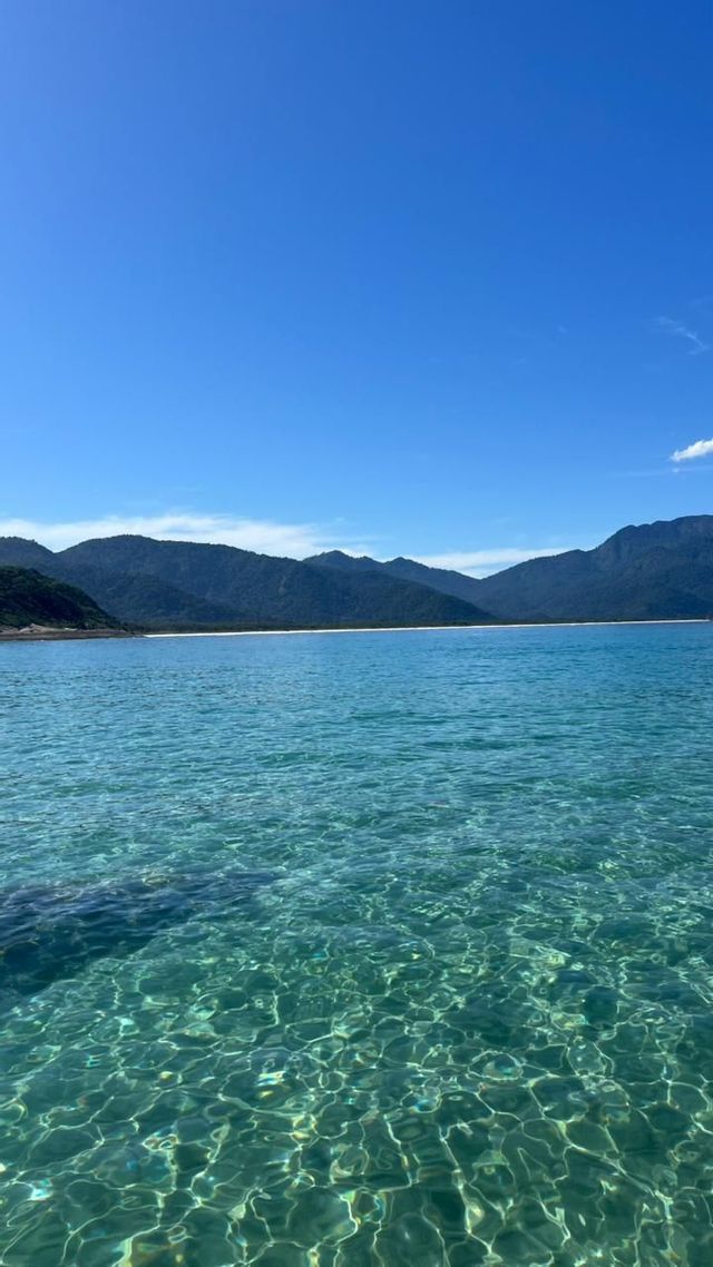 Sonnenlicht spiegelt sich auf klarem, ruhigem türkisfarbenem Wasser mit einer bergigen Küstenlinie im Hintergrund unter blauem Himmel.