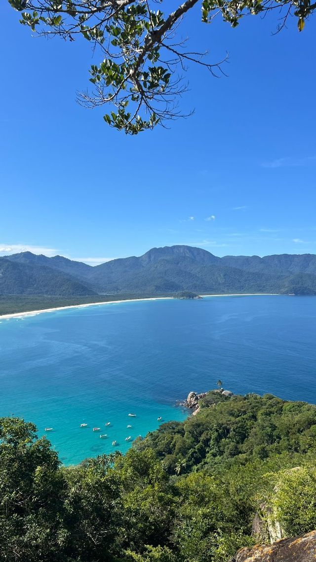 Vista dall'alto di una baia tropicale, con barche su acqua turchese, una spiaggia di sabbia bianca e montagne lontane sotto un cielo azzurro.