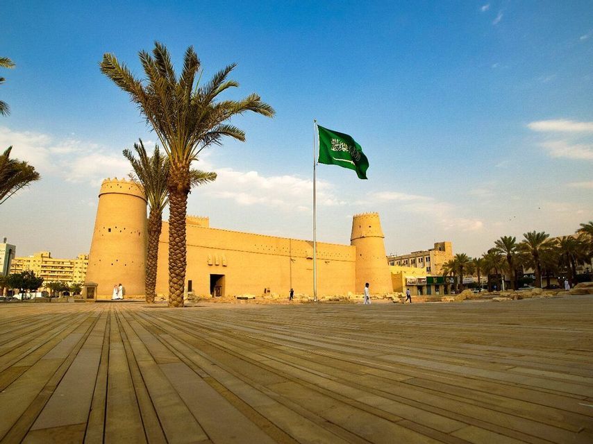 Le drapeau de l'Arabie Saoudite flotte sur un grand mât devant une forteresse couleur sable, avec des palmiers sur une grande place.