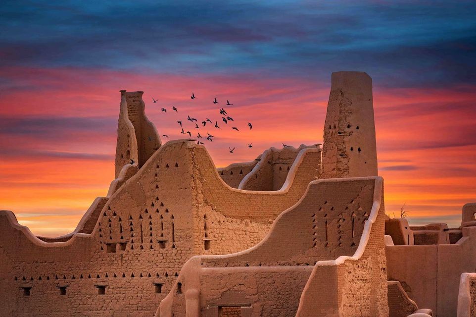 Un vol d'oiseaux survole d'anciennes ruines en briques de terre avec des tours, sous un ciel de coucher de soleil coloré.