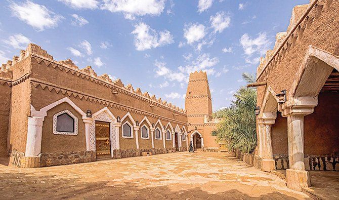 Un bâtiment traditionnel en pisé avec une tour et des arches blanches décoratives se dresse dans une cour en pierre sous un ciel bleu.