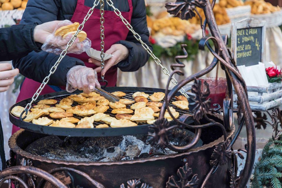 Un vendedor con guantes usa pinzas para asar pequeños trozos de queso en una parrilla circular grande suspendida sobre brasas calientes en un puesto de mercado.