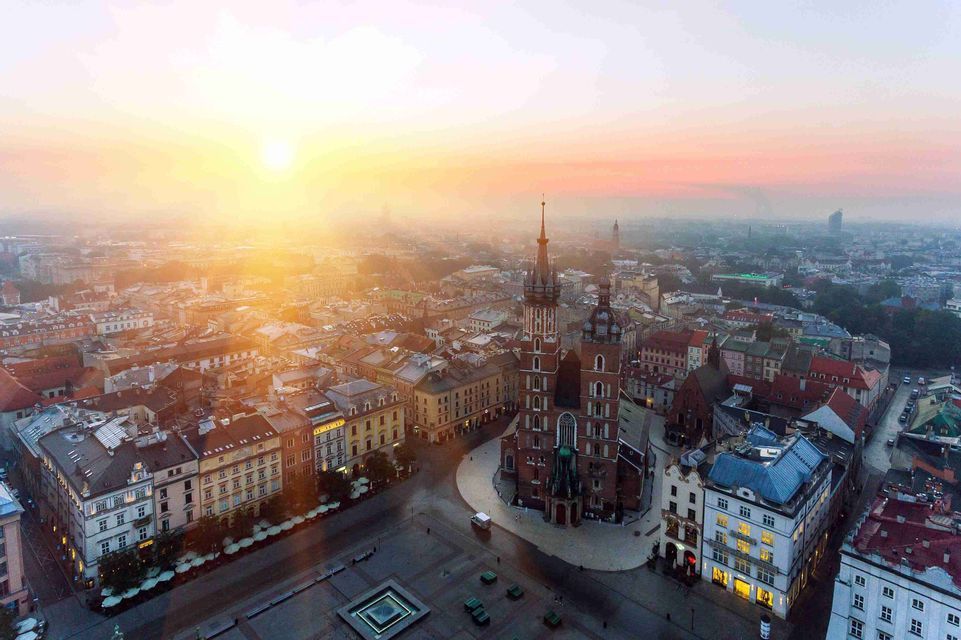 Una vista aérea de una plaza histórica y una gran iglesia de ladrillo bañadas por la luz dorada de un amanecer brumoso.