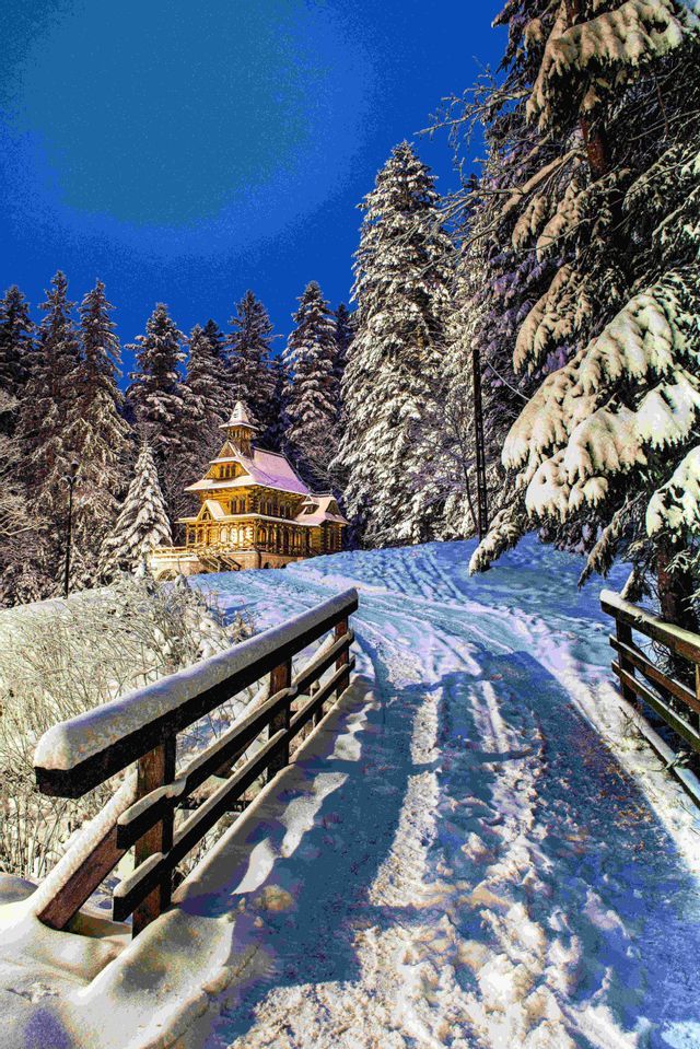 Una capilla de madera, iluminada por la noche, se asienta en un bosque nevado de pinos, con un puente cubierto de nieve en primer plano.