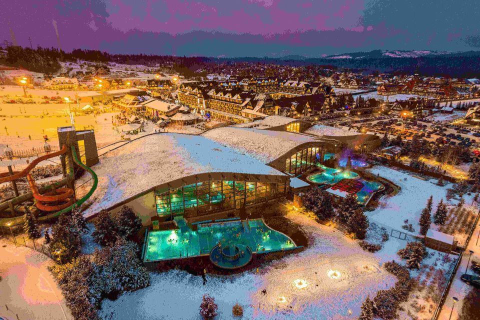Una vista aérea de un gran complejo de spa con piscinas turquesa iluminadas, situado en un pueblo cubierto de nieve al atardecer.