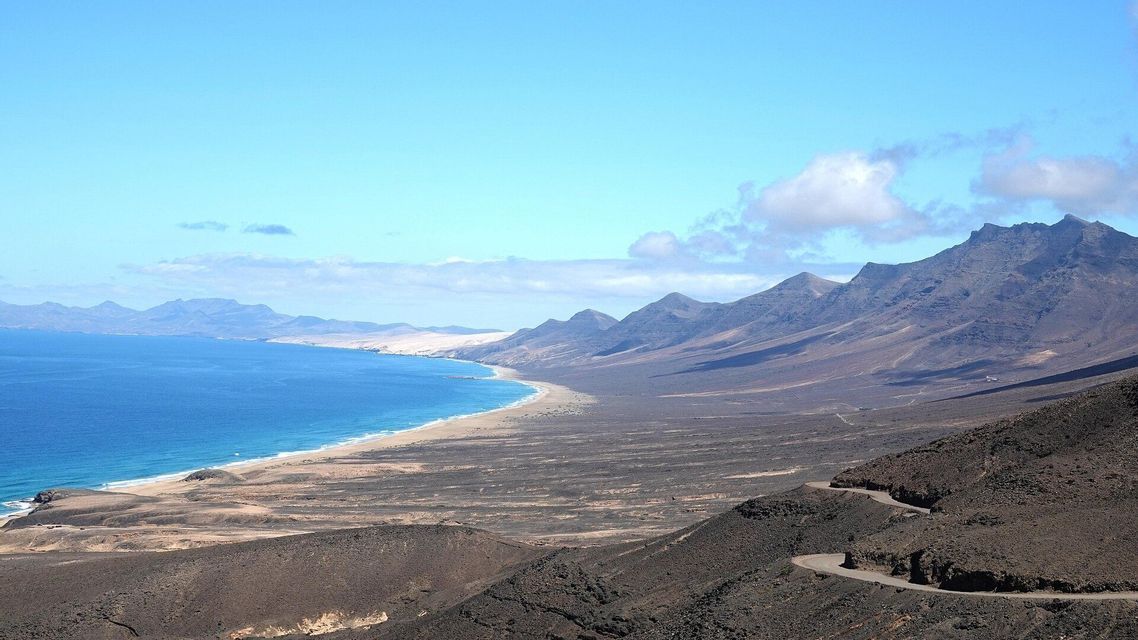 Vista aérea de una costa volcánica con una larga playa de arena que se une al océano azul, y una carretera sinuosa en primer plano.