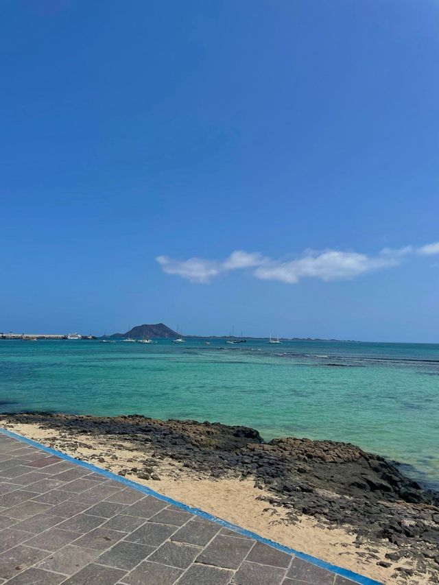 Un paseo pavimentado junto a una costa rocosa con vistas a una bahía turquesa con veleros y una isla lejana bajo un cielo azul claro.