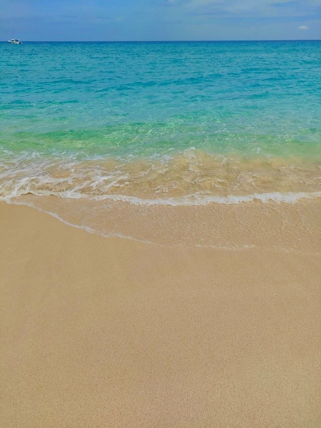 Una suave ola de agua turquesa cristalina baña una playa de arena bajo un cielo azul, con un pequeño bote en el horizonte.