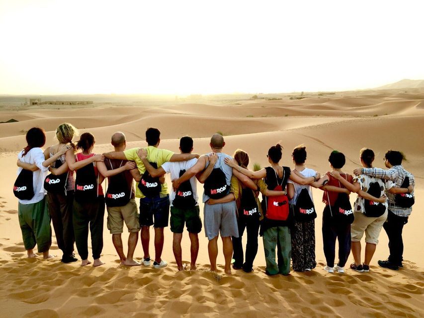 A WeRoad group trip standing arm-in-arm on a sand dune, looking out over a vast desert landscape at sunset.