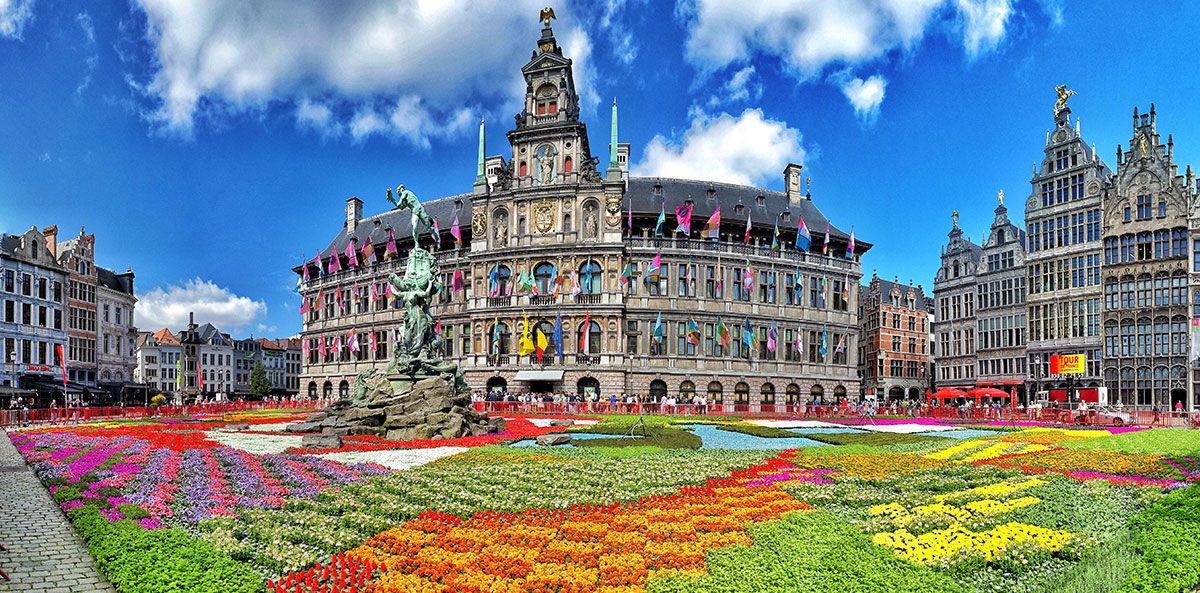 Une vaste étendue de fleurs colorées recouvre une place historique devant un bâtiment orné de nombreux drapeaux.