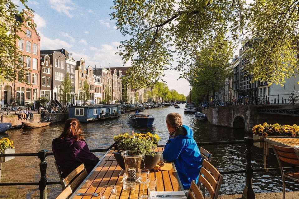 Deux personnes assises à une table en bois sur une terrasse, regardant un canal de la ville avec des bateaux et des bâtiments.