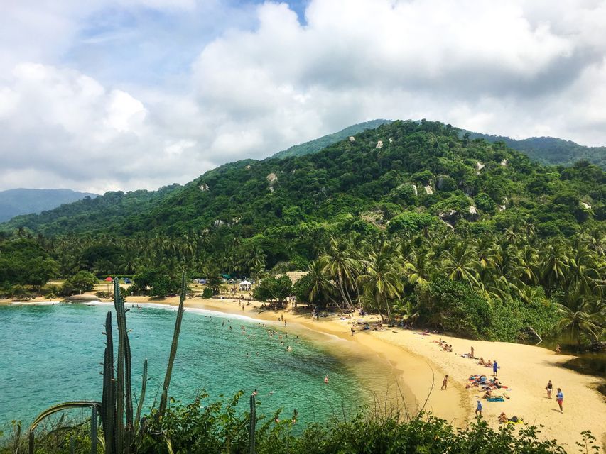 Vista dall'alto di una spiaggia tropicale dove le persone nuotano in acque turchesi, con una grande collina boscosa sullo sfondo.