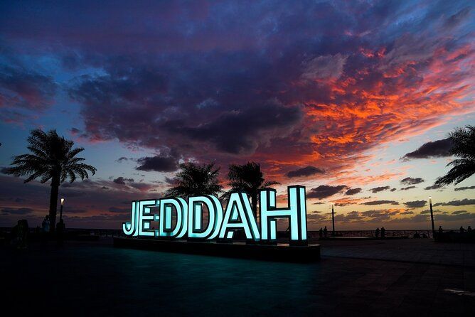 Une enseigne lumineuse épelant 'JEDDAH' sur un front de mer avec des palmiers sous un ciel de coucher de soleil coloré.