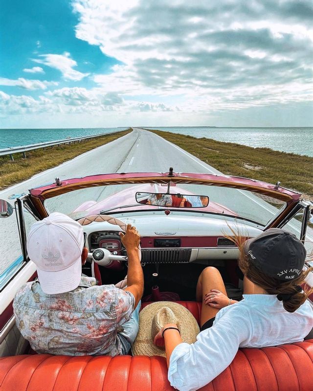 Due membri di un gruppo WeRoad a bordo di una decappottabile rosa d'epoca, che guidano su una strada costiera lungo il mare.