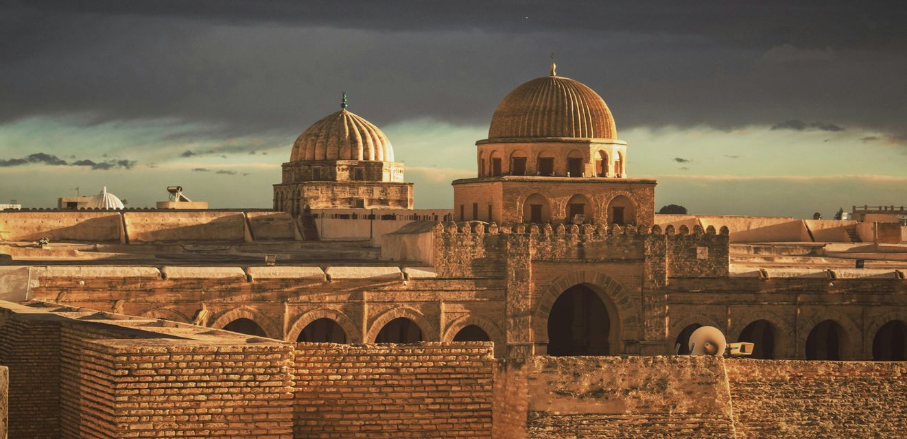 Ancient stone city rooftops and domes illuminated by golden sunlight under a dark, stormy sky.