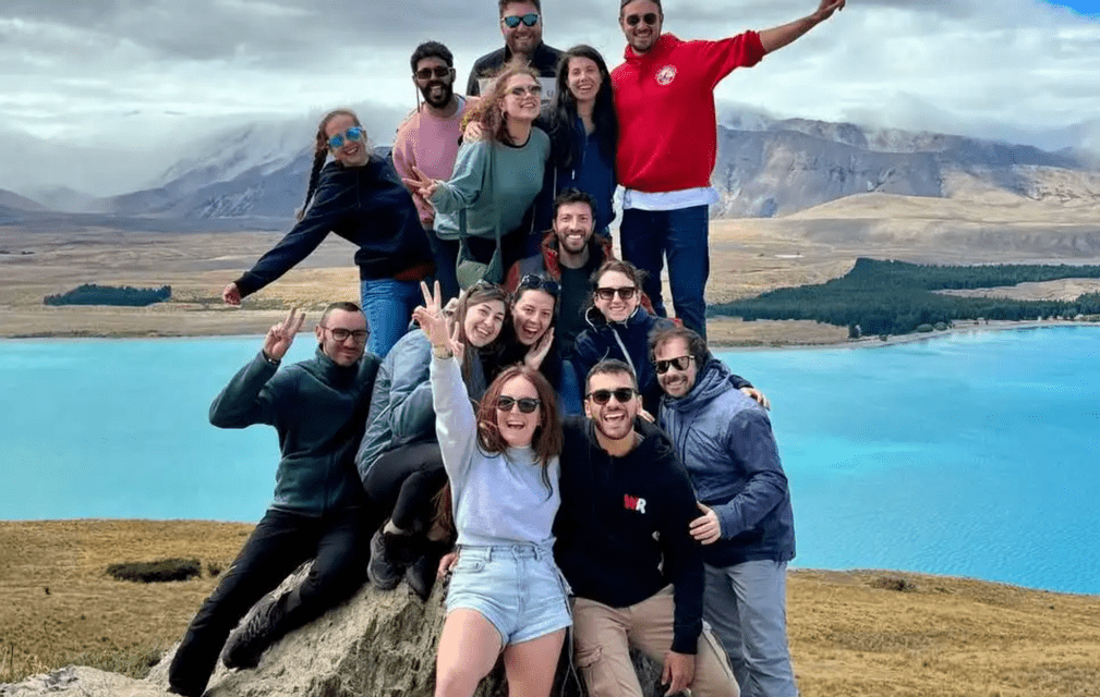 A WeRoad group trip poses for a photo on a rocky outcrop, overlooking a turquoise lake and distant mountains.