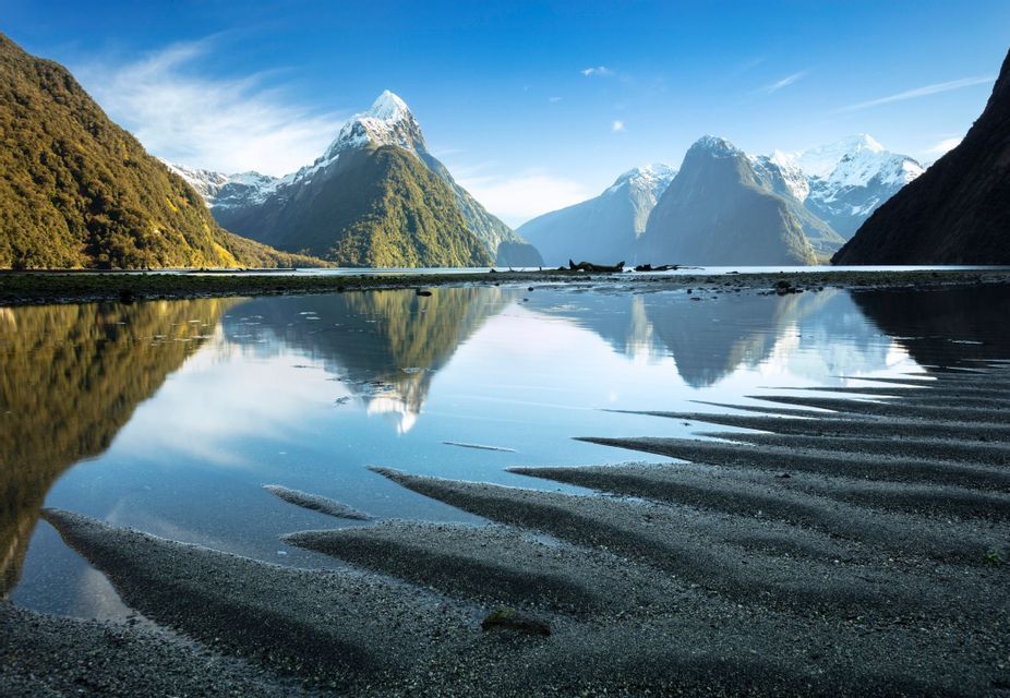 Snow-capped mountains and green hills are reflected in calm fjord waters, with rippled wet sand in the foreground.