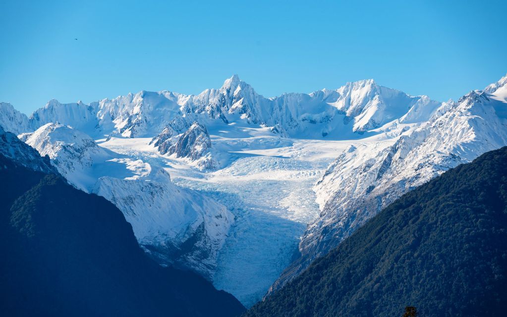 A large glacier flows down from a mountain range of snow-covered peaks, framed by dark forested slopes under a clear blue sky.
