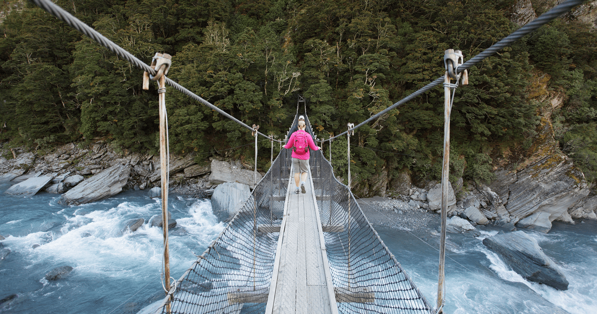 A person wearing a pink backpack walks across a narrow suspension bridge over a rushing river in a forested canyon.