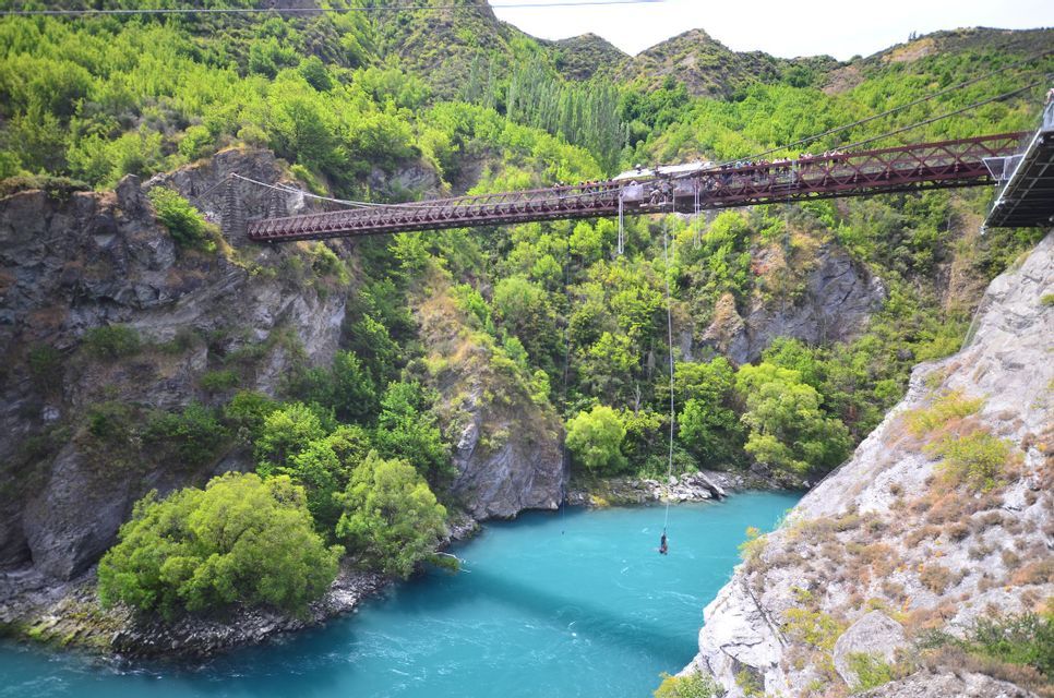A person bungee jumping from a bridge suspended over a turquoise river in a lush green canyon.