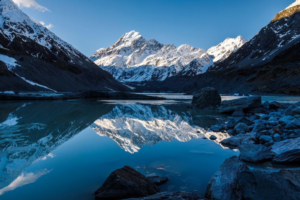 A snow-capped mountain range is perfectly reflected in the still water of a calm lake under a clear blue sky.