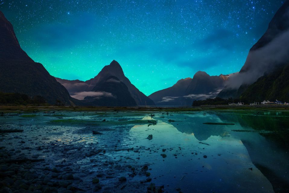 Silhouetted mountains are reflected in the water of a fjord under a vibrant green and blue starry night sky.