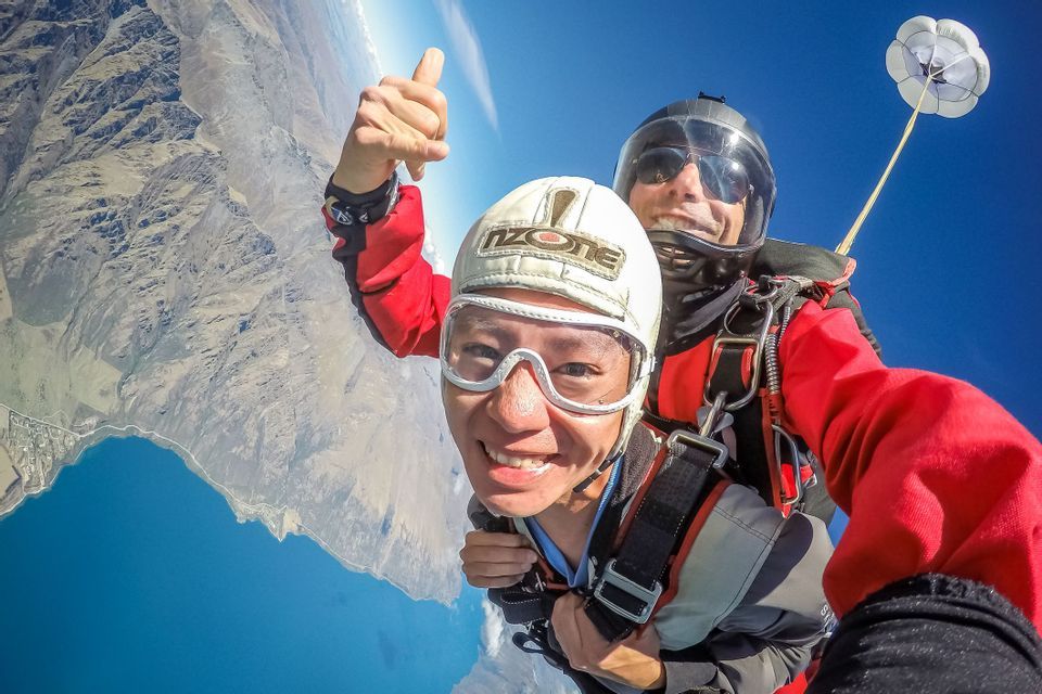 Two people take a selfie during a tandem skydive, smiling high above a mountainous landscape and a large blue lake.