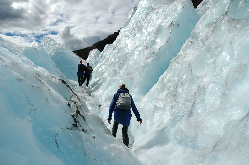 A WeRoad group trip trekking single file through a narrow crevasse in a large, blue-tinged glacier under a cloudy sky.