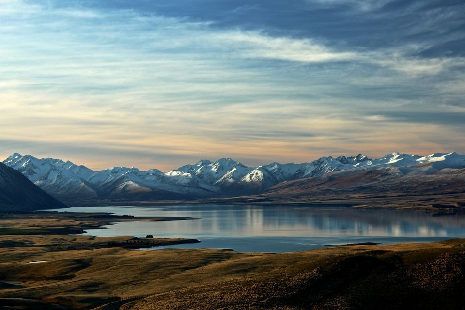 A calm lake reflects a range of snow-capped mountains under a colorful sky, with grassy hills in the foreground.