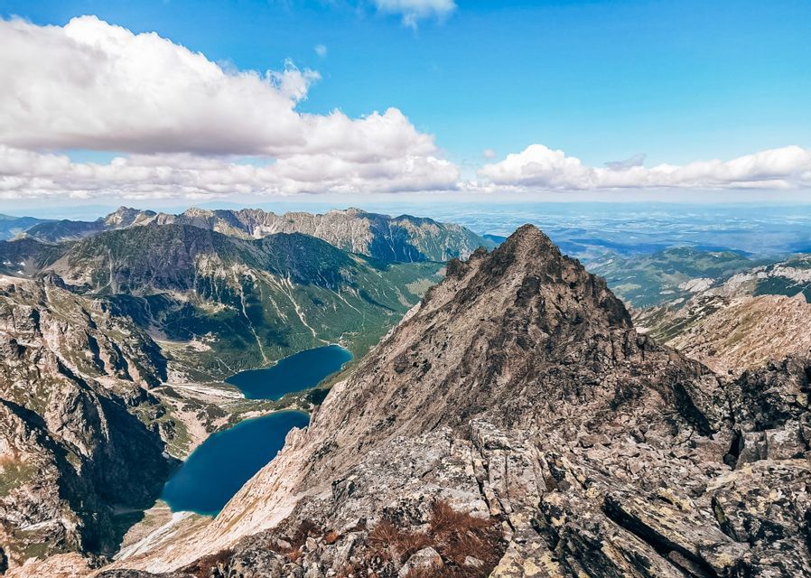 A panoramic view from a rocky mountain peak overlooking a vast mountain range with two blue alpine lakes in a valley below.